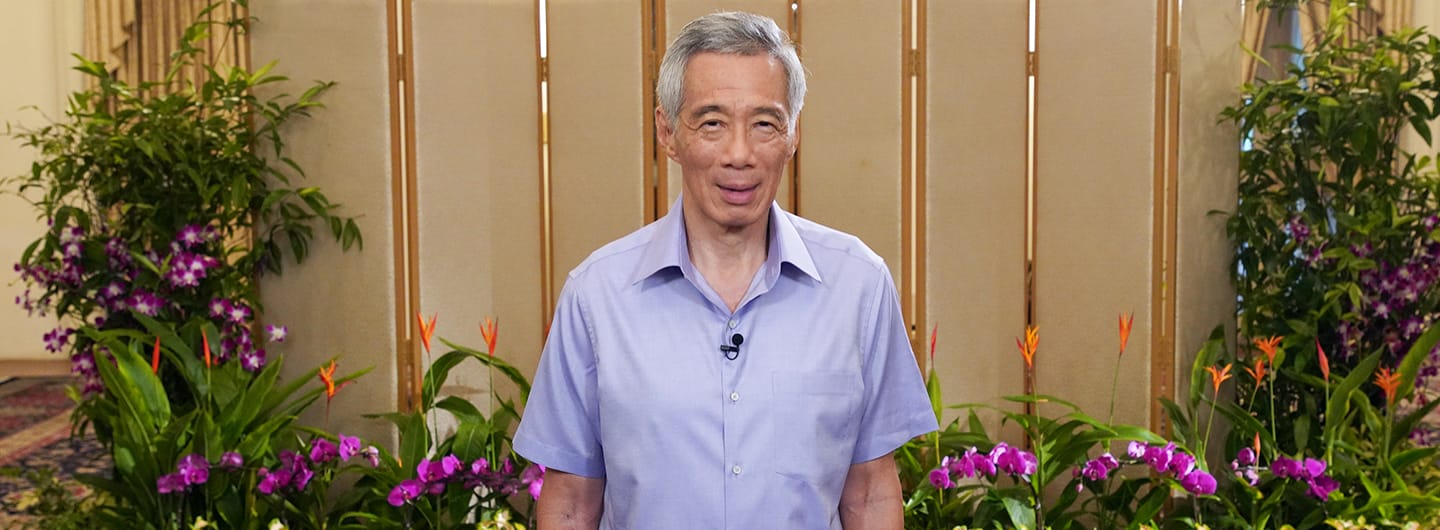 Lee Hsien Loong in a pale blue shirt stands before a patterned screen flanked by orchids.